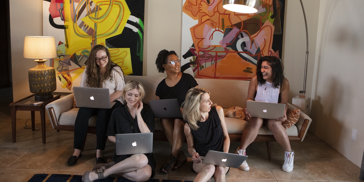 Rachel, Laneia, Carmen, Riese and Sarah all sit together near a couch on their laptops. Sarah's laughing, Carmen and Riese are looking at her, and Rachel and Laneia are looking at their computers.