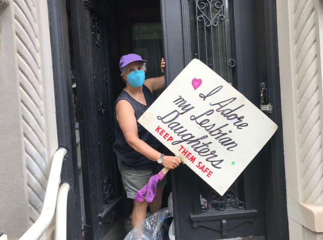 Frances Goldin in a mask at her front porch holding the sign "I love my lesbian daughters. Keep them safe."