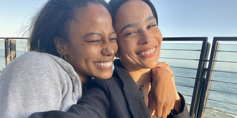 Zoe Kravitz and Taylour Paige are smiling together in the sunset against blue water and equally blue skies. Taylour's hands are draped over Zoe's shoulders.