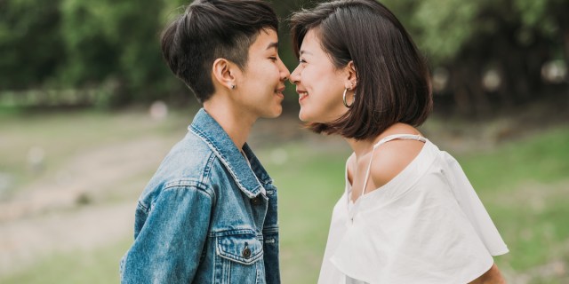 A person in a denim jacket with short, partially shaved hair touches noses with a person in a white shirt with slightly longer hair