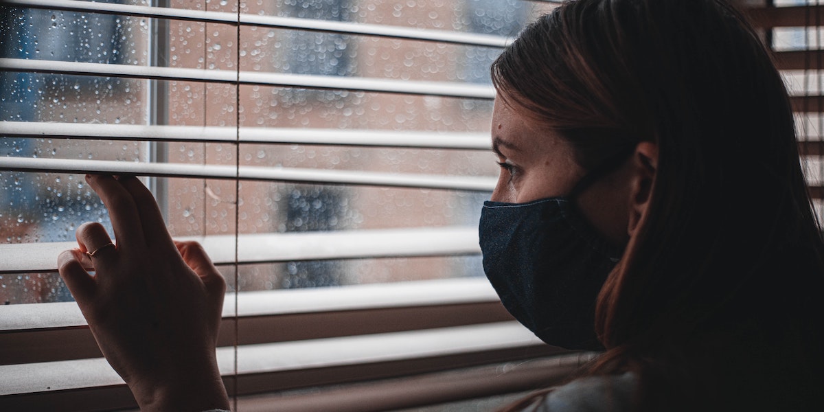 A girl wearing a facemask inside a dimly-lit indoor space lifts open the space between two slats of window blinds to tentatively peer out