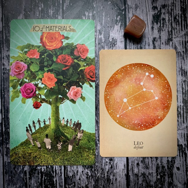 A tarot card for 10 of Materials featuring a group of people circled around a blooming tree; a constellation card reading Leo: Defeat, and a brown crystal