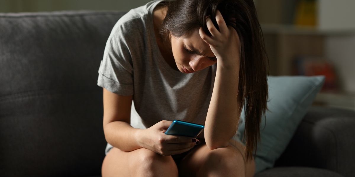A distraught-looking teen girl sits on a couch, looking at her phone screen with concern as she runs her hands through her long hair.