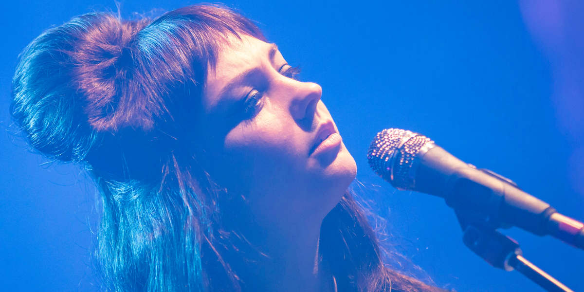 Angel Olsen, who came out as gay today, with long brunette hair and bangs, is photographed on stage, surrounded by blue lighting. There's a microphone in front of her.
