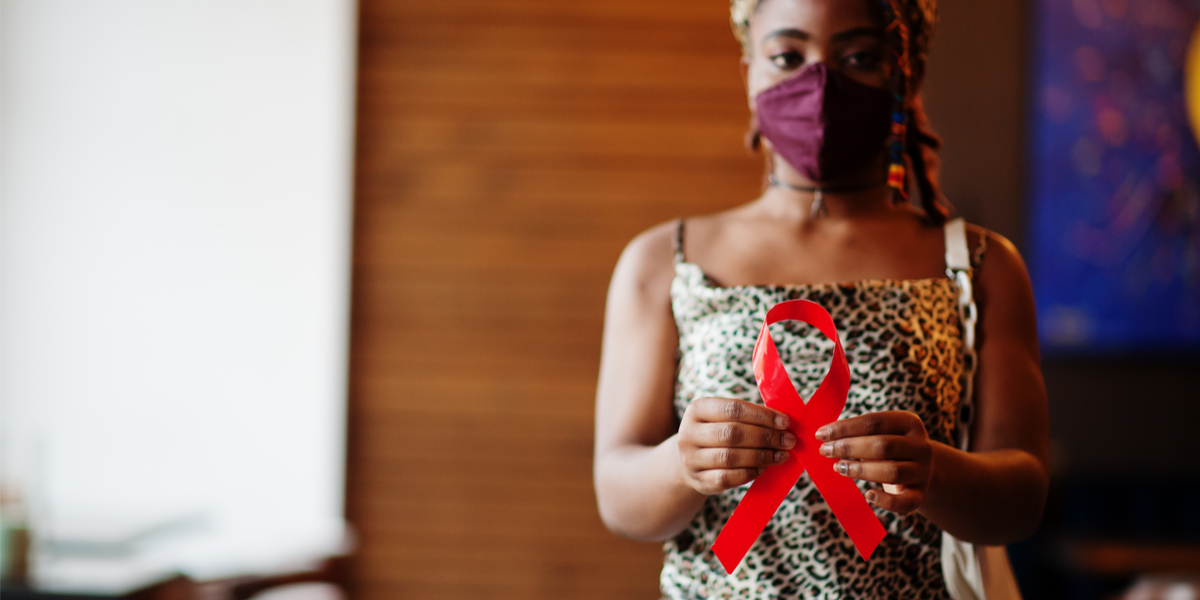 A young Black woman wearing a face mask holds a red ribbon out in front of her body.