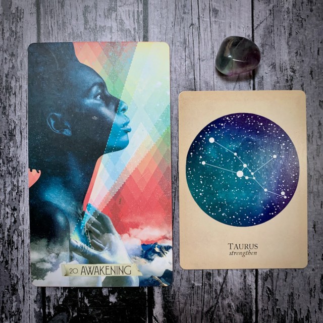 A photo of the tarot card Awakening, which features a Black woman's face in profile, the constellation card that read Taurus: Strengthen, and a grey crystal