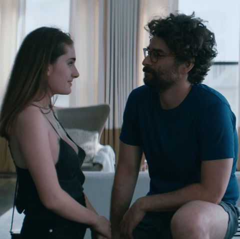 Image shows a woman in a black tank top talking to a man in glasses and tshirt in a living room.