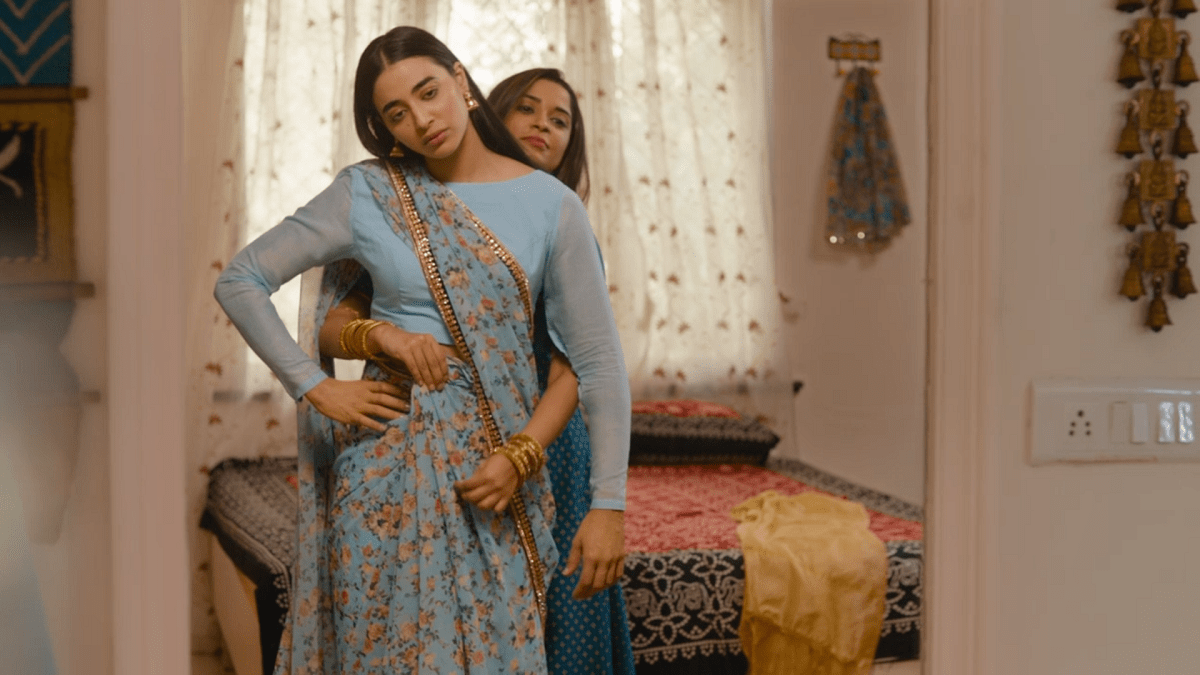 Umang's sister-in-law and ex, Pinky helps Umang get dressed in a pale blue sari. Pinky stands behind Umang setting the pleats in place, while Umang looks dejected.