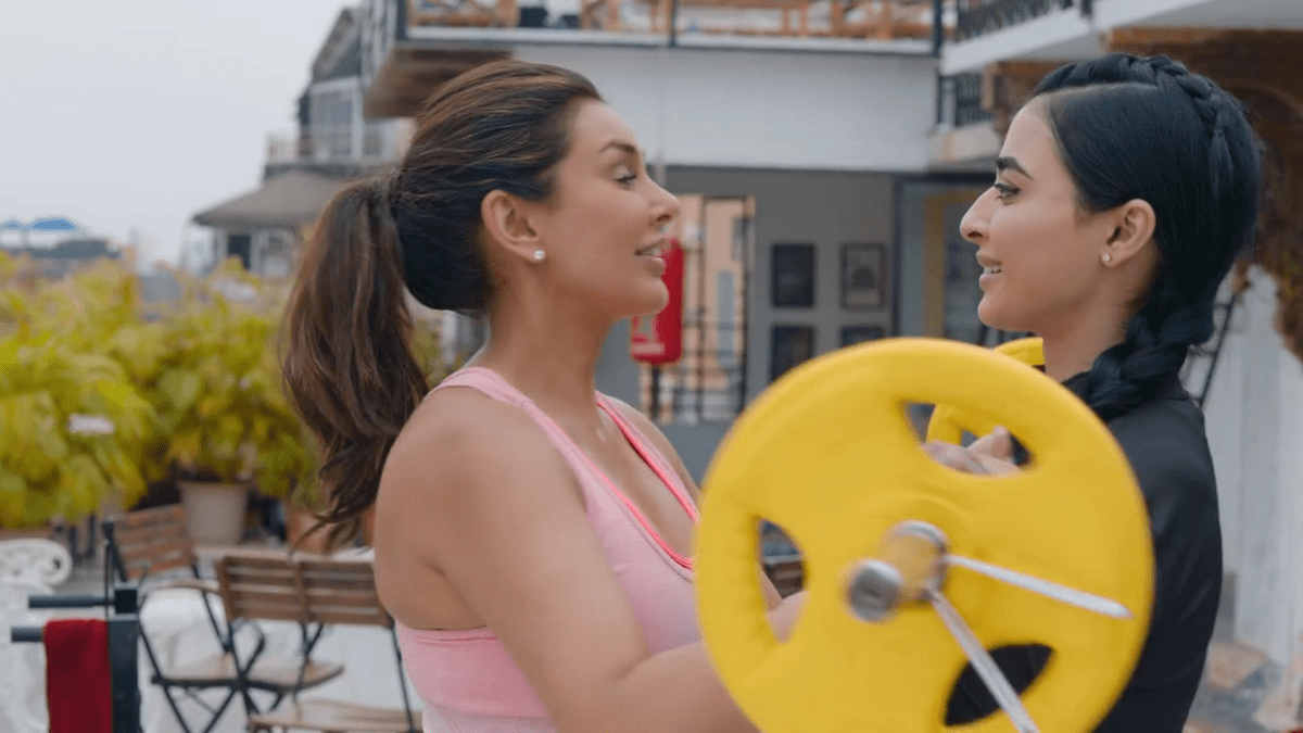 Standing on a rooftop in a peach-colored workout top, Lisa Ray, playing the character of Samara Kapoor hands a barbell to Umang, played by actress Bani J. 