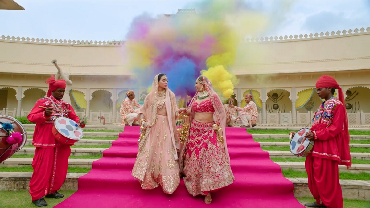 Dressed in pink lenghas, traditional South Asian wedding attire, Umang and Samara walk down a red carpet. Behind them is a colorful cloud and alongside their path are men dressed in red outfits playing drums with hearts on them. In the background we see the fort where the wedding is set.