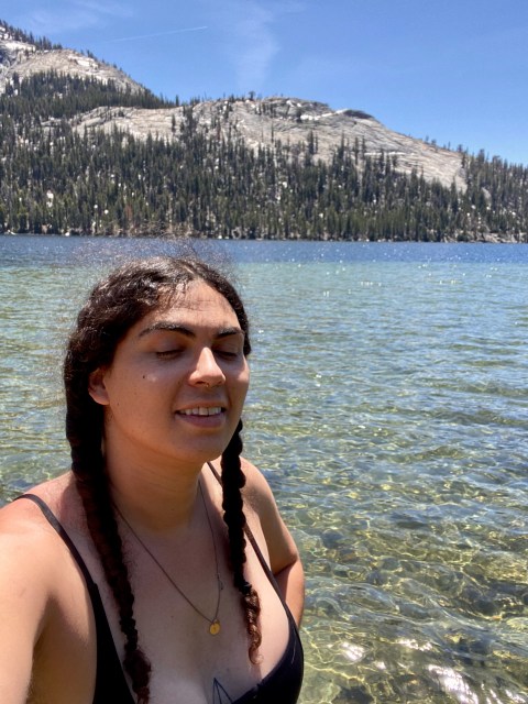 Abeni, a queer person in swimwear, stands in water in front of mountains in a black swimsuit