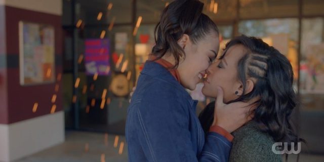 Screenshot from Legacies: Bisexual witch Josie smiles as she goes in to kiss her queer werewolf gal pal as sparks literally fly behind them.