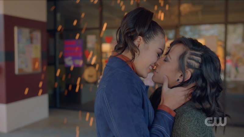 Screenshot from Legacies: Bisexual witch Josie smiles as she goes in to kiss her queer werewolf gal pal as sparks literally fly behind them.