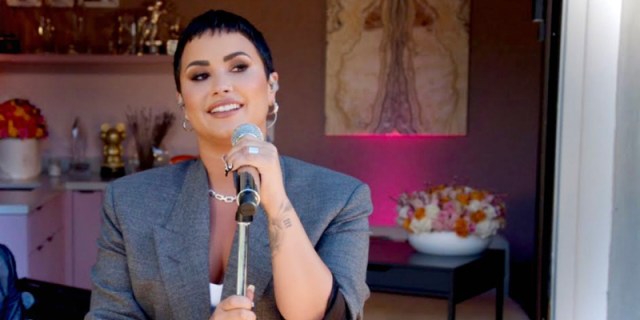 Demi Lovato in their new signature short hair cut is in a oversized grey blazer performing in front a microphone, the wall has a pink background