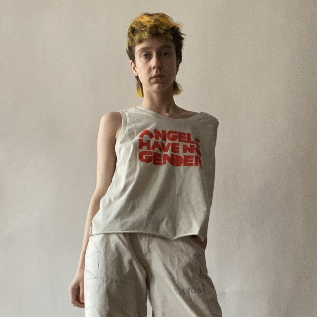 Model in beige pants and a sleeveless beige tee that reads ANGELS HAVE NO GENDER in red