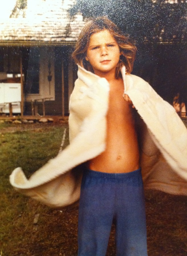 Daniela at 7 stands in front of a house in swim trunks with a towel around them