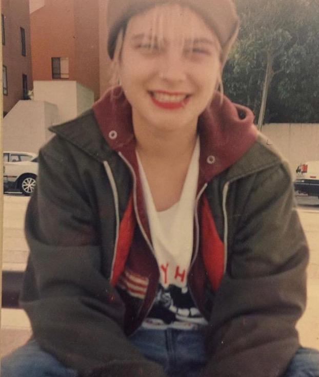 Daniela at 18 is wear a hoodie and a jacket and a beanie. They're wearing red lipstick.