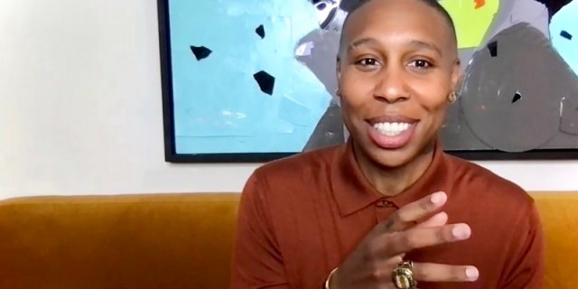 In an interview with Autostraddle via zoom, Lena Waithe is in a brown sweater and wearing many gold rings across her fingers. She's on a brown couch with light blue artwork behind her against a white wall. She's smiling and talking with her hands.