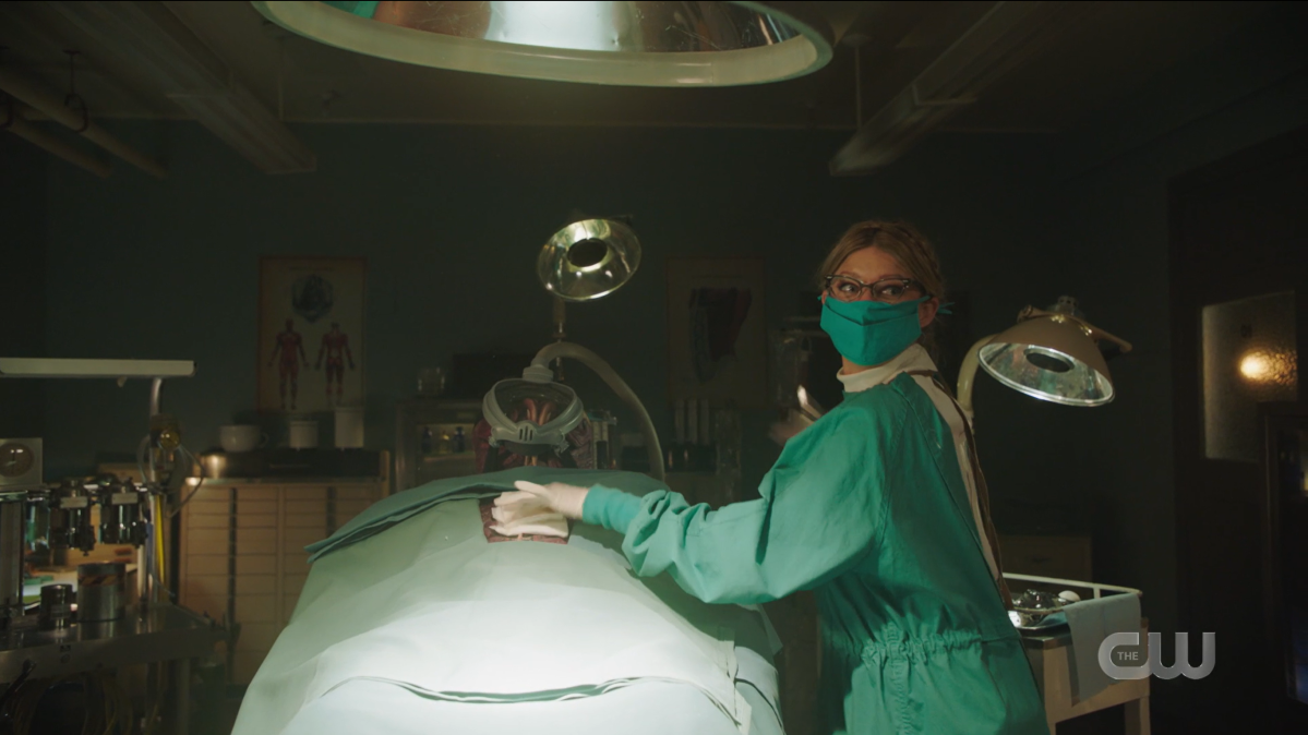 Ava is in scrubs doing surgery on an alien and looking mischievous.