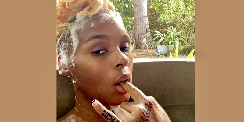 Janelle Monae is in a hot tub with suds in her hair. She is sucking on her finger.