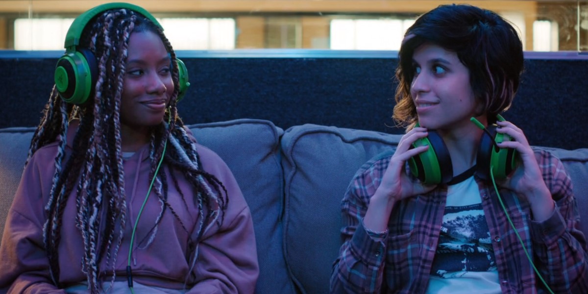 Mythic Quest queer gamers: Dana and Rachel sit on a couch with gamer headphones on smiling at each other.