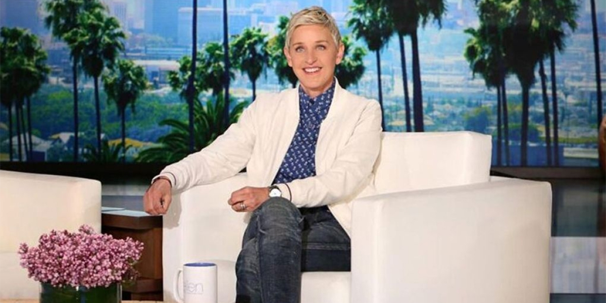 Ellen DeGeneres sits on the set of her talkshow in a white blazer, blue button-up, and jeans