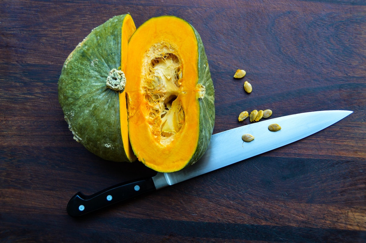An overhead photo of a sliced kabocha squash with a chef's knife, with some of the seeds spilling onto the table.