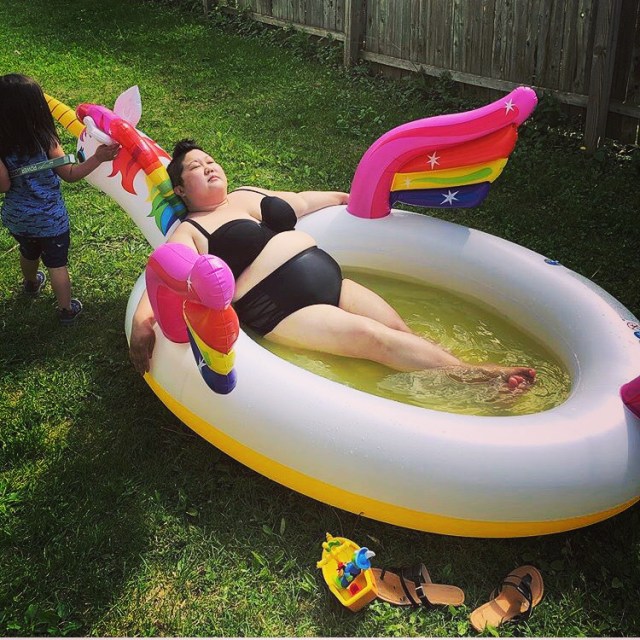 Kaeyln relaxes in an inflatable unicorn-shaped pool while wearing a black bikini and black bikini bottoms. A child stands near the pool.