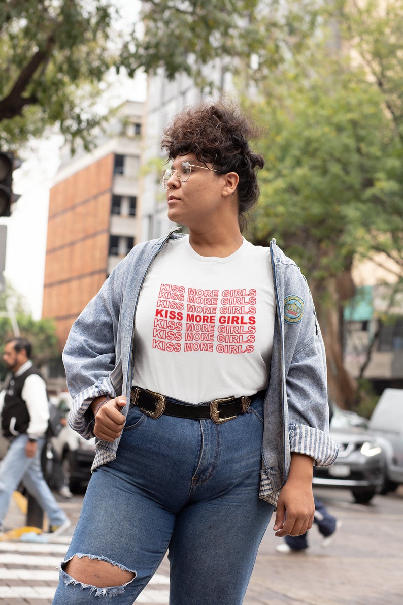 Model in jeans on a busy city street, the t-shirt is white and says KISS MORE GIRLS over and over like on the shopping bags that have the red print that say the same thing over and over