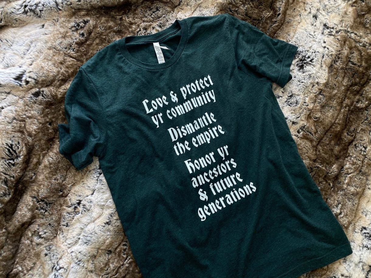 Dark green t-shirt that says 'Love & protect yr community / dismantle the empire / honor yr ancestors and future generations" on it in white Old English style lettering