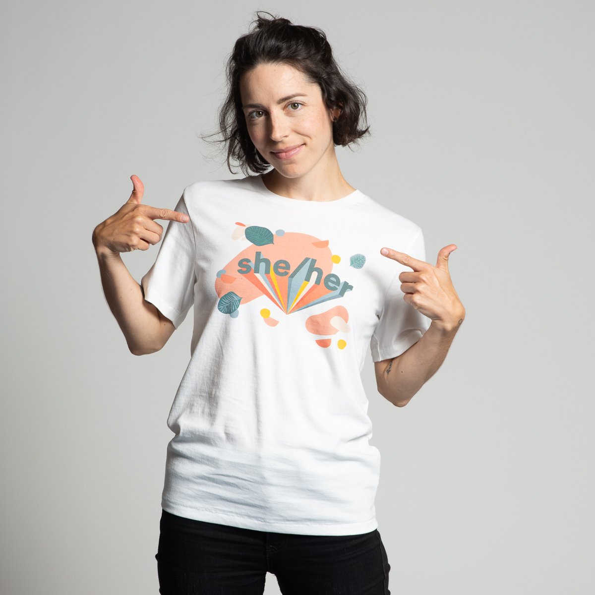 Model wearing a white tee with "she/her" on it in peach, teal, yellow and similar colors in a fan pattern