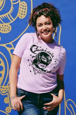 Smiling model in front of a blue wall, wearing a lavender t-shirt that says "Luchadora" in script above an illustration a woman in a mask.