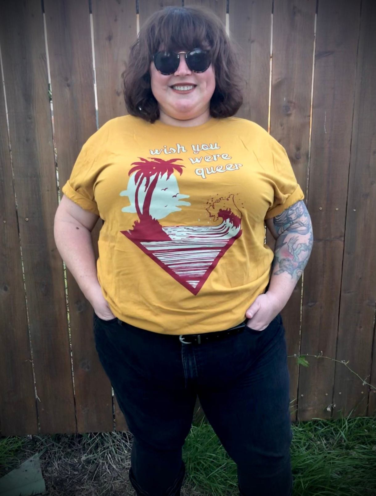 Model wearing a mustard yellow t-shirt that says "wish you were queer" on it with a picture of a palm tree on an island and a wave in maroon and light blue.