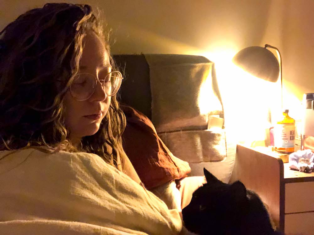 A dimly lit photo of Rachel sitting up in bed, with a nightstand visible behind her and a small black cat visible in the foreground