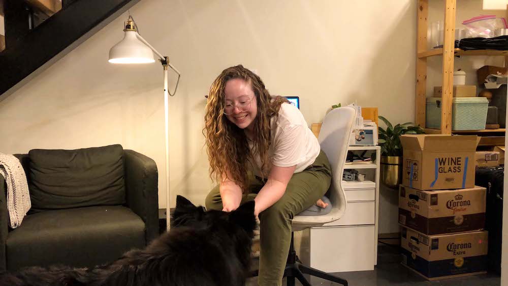 Rachel is seated at her desk next to an armchair and pile of unpacked moving boxes, smiling and leaning forward to greet a fluffy black dog on the ground in front of her.