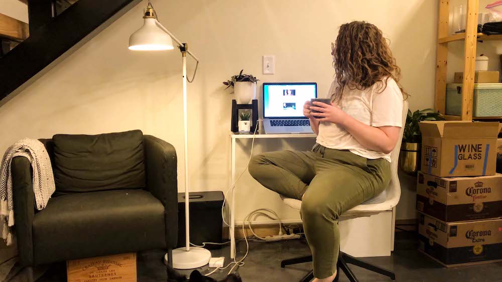 Rachel, a white woman with curly hair wearing green sweatpants and a white t-shirt, is sitting at a small white desk next to a lamp, armchair and a pile of unpacked moving boxes. She's turned to the side, cupping a cup of coffee while looking at her laptop screen.