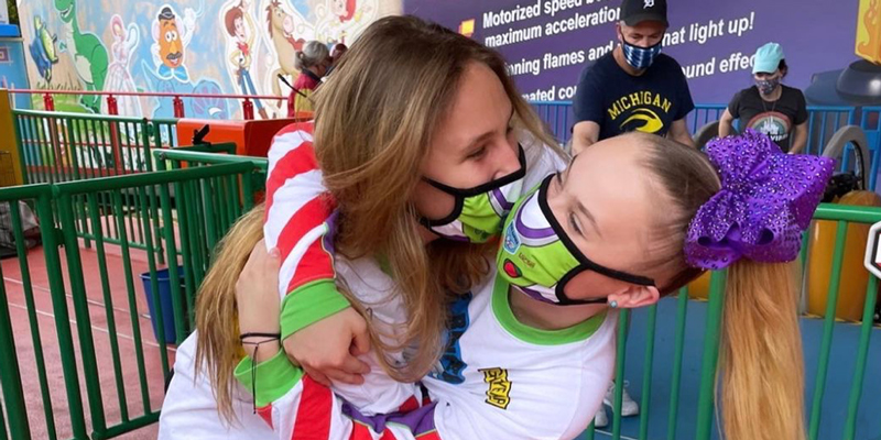 Jojo Siwa is in a Toy Story themed face mask and is kissing her girlfriend in an amusement park (who is also wearing a Toy Story face mask)