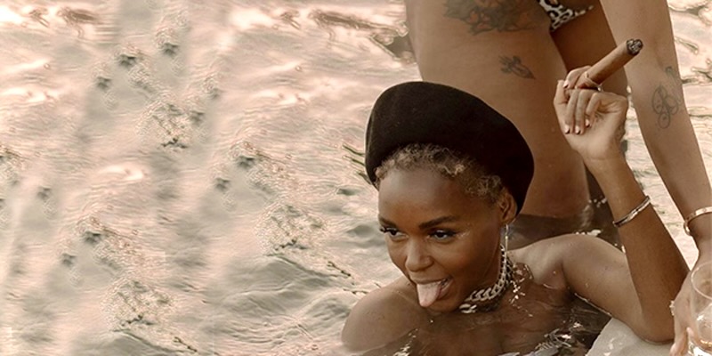 Janelle Monáe is in a pool at sunset. She s smoking a cigar and sticking her tongue out while smiling, there's a woman in a bikini getting out of the pool behind her.