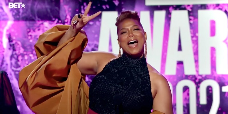 Queen Latifah says Happy Pride and calls her partner "my love" from the 2021 BET Awards Show. Queen Latifah is dressed up in a Black dress with a gold shawl. She smiles and flashes a peace sign. She's in front of a purple backdrop from the BET Awards Stage.