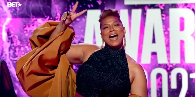 Queen Latifah says Happy Pride and calls her partner "my love" from the 2021 BET Awards Show. Queen Latifah is dressed up in a Black dress with a gold shawl. She smiles and flashes a peace sign. She's in front of a purple backdrop from the BET Awards Stage.