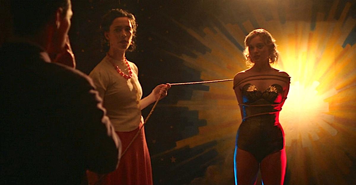 A man watches on as one woman holds onto a rope that's tied around a younger woman who is dressed like Wonder Woman.