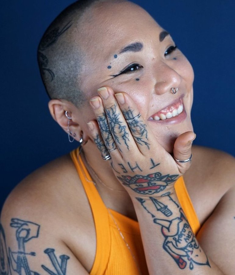 A person smiles against a dark blue background, showing off several rings, earrings, and nose rings.
