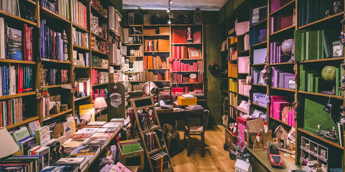 A grainy photo of a corner of a small bookstore in warm lighting, with shelves full of colorful books and a small stepladder and desk