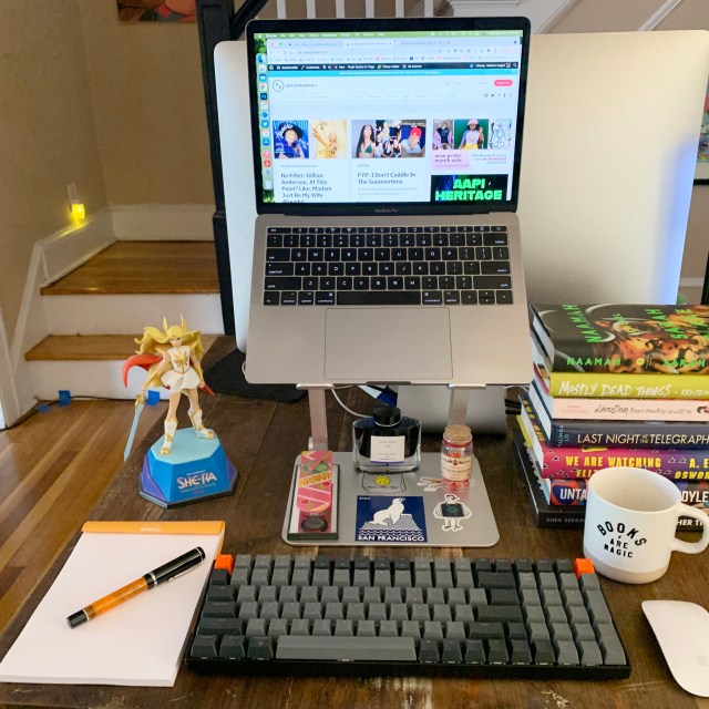 This is Heather Hogan's Desk! It has a laptop with separate keyboard and mouse, notepad and pen, She-Ra figurine, mug, pile of books