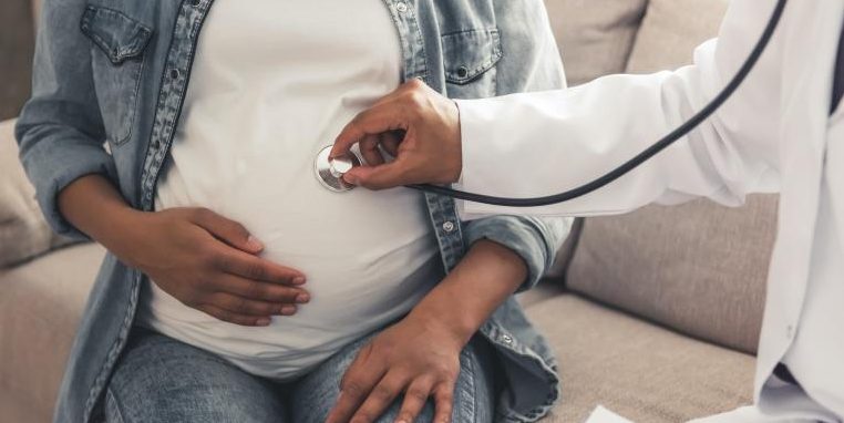 A photo of a brown-skinned pregnant person seated on a sofa with one hand cradling their belly while a person in a white lab coat, also brown-skinned, holds a stethoscope over their belly.