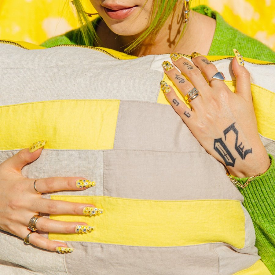 A person with green hair and long colorful bedazzled nails holds a grey and yellow pillow.