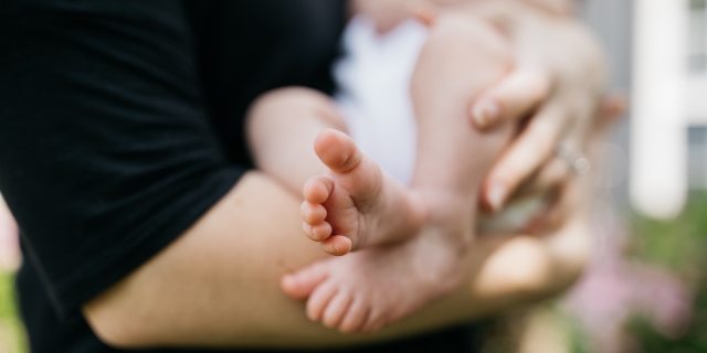 A close-up portrait of someone cradling a very young baby, with the person's arms visible and the baby's bare feet up close to the lens.