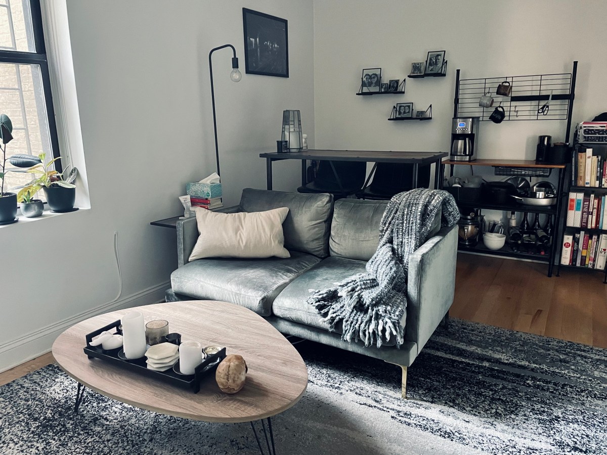 Alternate angle of the living room, showing the front of the couch and the coffee table. Behind the couch the bar table and baker's rack are visible, along with shelves and photos.