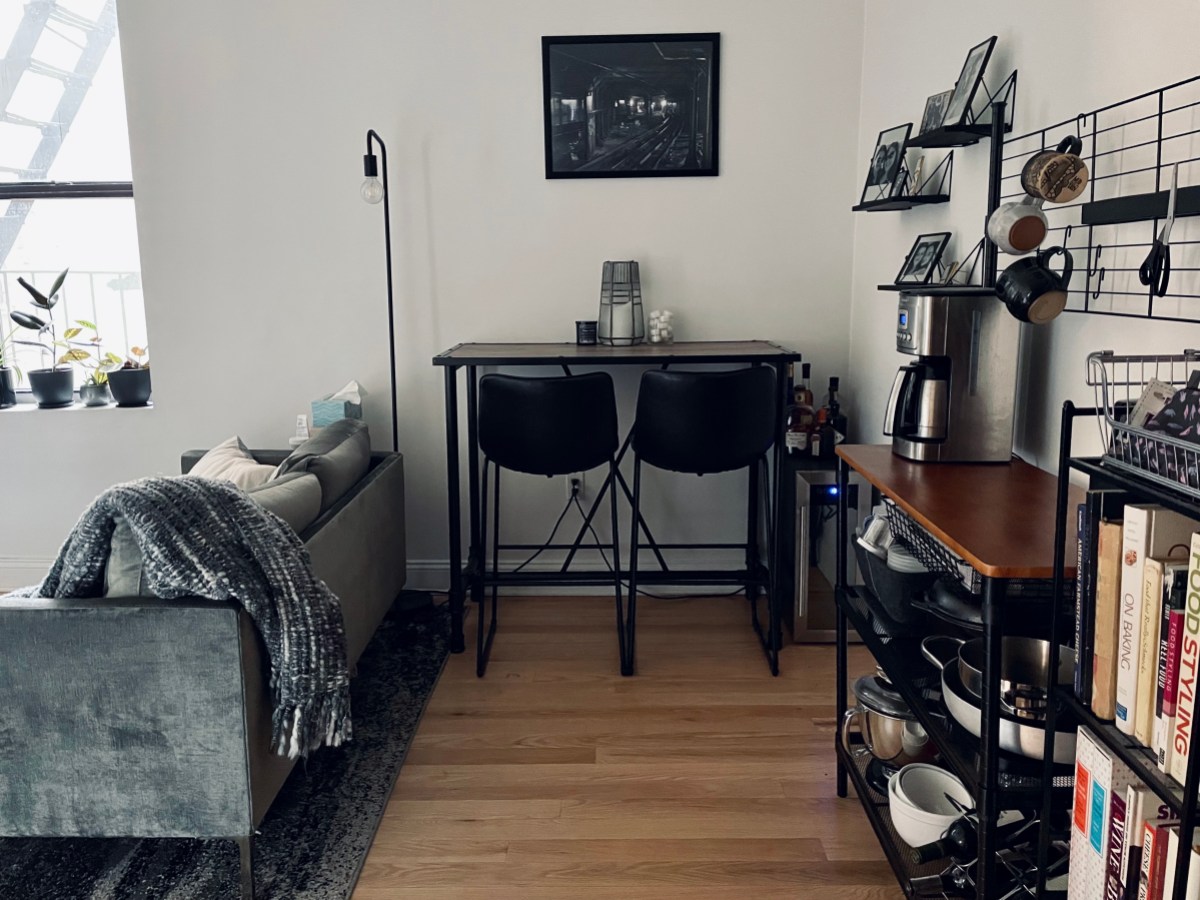 Image of one corner of a living space, with a baker's rack and shelves against the right wall, a bar table with two bar stools against the back wall, and the back of a sofa against the left side.