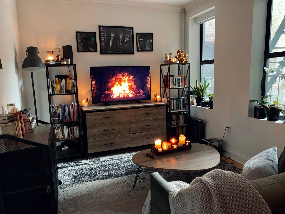 Living room with television playing fire video, bookcases covered in lit candles, and a coffee table in front of a sofa that also holds a tray of burning candles. Windows on the side have plants on the sills, a rug is on the floor, and the couch has a large throw blanket on one corner